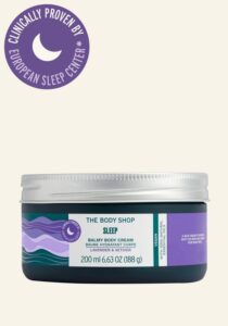 A short cylindrical clear glass jar full of some dark green liquid with a silver Aluminium Lid and a white label that has The Body Shop Sleep Balmy Body Cream written in dark green writing and some pictures of some light purple swirls on it, on a white background.