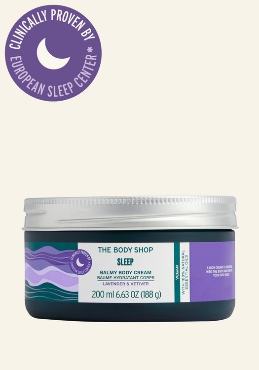 A short cylindrical clear glass jar full of some dark green liquid with a silver Aluminium Lid and a white label that has The Body Shop Sleep Balmy Body Cream written in dark green writing and some pictures of some light purple swirls on it, on a white background.