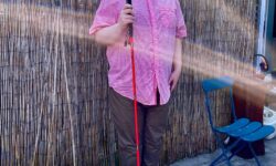 A tall bearded man wearing a pair of silver and black aviator sunglasses with some light green lenses, a bright orange short sleeve shirt that has a white grid pattern all over it, a dark red v neck t-shirt, a pair of dark brown cord trousers and a pair of shiny red shoes holding a bright red cane stood against a dark wooden fence.
