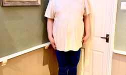 A tall bearded man wearing a bright white crew neck t-shirt, a pair of shiny dark blue leggings and a pair of black and white striped bamboo socks stood against a light grey wall.