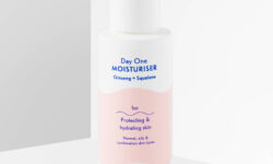 A tall cylindrical white plastic bottle with a white pump lid and a white label that has By Beauty Bay Day One Moisturiser with Ginseng and Squalane written in dark blue writing and a picture of a pale peach stripe on it, on a white background.
