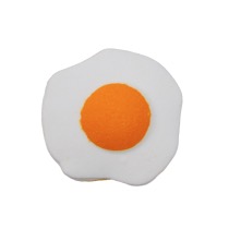 A large bright white fried egg shaped bath Bomb that has a large bright golden yellow yolk in the middle of it, on a white background.
