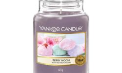 A tall cylindrical glass jar full of some dark purple wax with a label that has Yankee Candle written in white writing, Berry Mochi written in black writing, and some pictures of some bright pink, blue and purple balls of Mochi on it, on a white background.