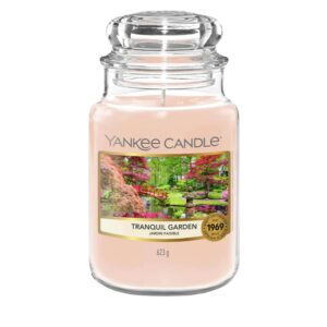 A tall cylindrical glass jar full of some pale Pink wax with a label that has Yankee Candle written in white writing, Tranquil Garden written in black writing, and a picture of a Japanese Garden on it, on a white background.