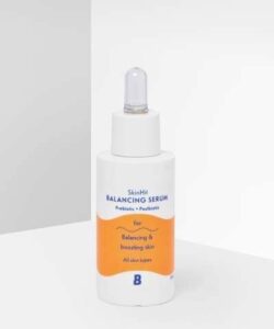 A short cylindrical white plastic bottle with a white rubber pipette lid that has By Beauty Bay SkinHit Balancing Serum with Prebiotic and Postbiotic written inn dark blue writing and a picture of a bright orange stripe on it, on a white background.