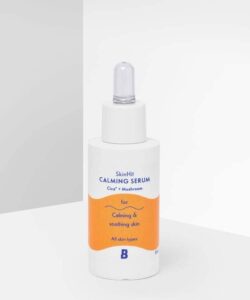 A short cylindrical white plastic bottle with a white rubber pipette lid that has By Beauty Bay SkinHit Calming Serum with Cica and Mushroom written inn dark blue writing and a picture of a bright orange stripe on it, on a white background.
