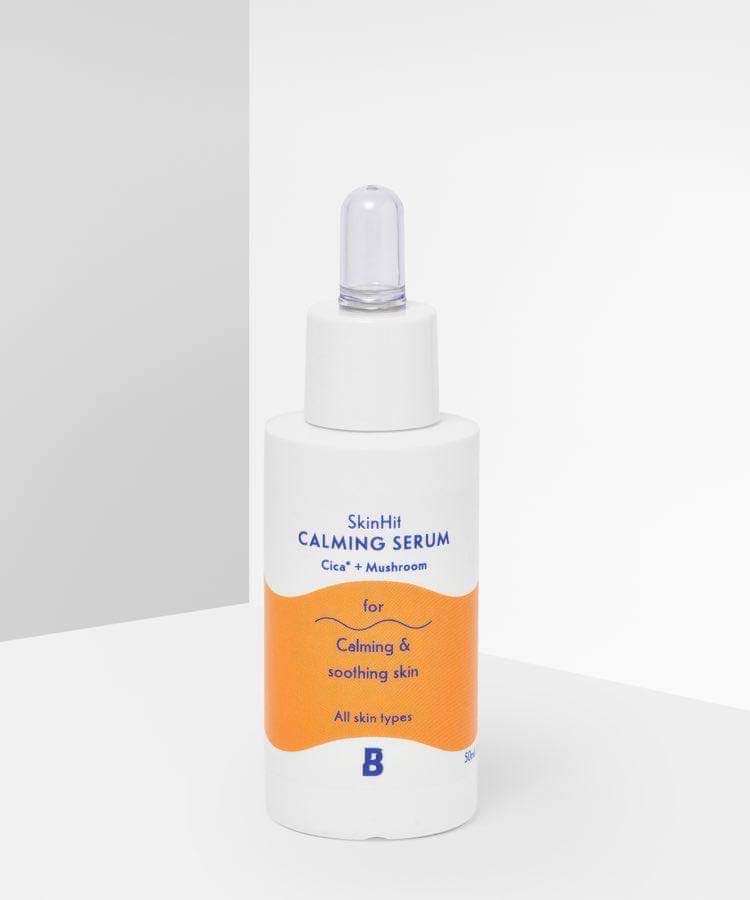 A short cylindrical white plastic bottle with a white rubber pipette lid that has By Beauty Bay SkinHit Calming Serum with Cica and Mushroom written inn dark blue writing and a picture of a bright orange stripe on it, on a white background.