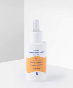 A short cylindrical white plastic bottle with a white rubber pipette lid that has By Beauty Bay SkinHit Correcting Serum with AHA and PHA written inn dark blue writing and a picture of a bright orange stripe on it, on a white background.