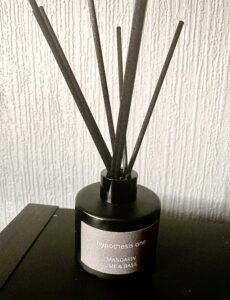 A short cylindrical black glass jar with a light grey label that has Hypothesis One Mandarin, Lime & Basil Intense Fragrance Diffuser written in bold white writing on it and some black wooden reeds coming out of the top of it on top of a black glossy wooden cabinet.