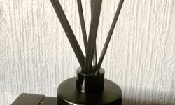 A short cylindrical black glass jar with a light grey label that has Hypothesis One Mandarin, Lime & Basil Intense Fragrance Diffuser written in bold white writing on it and some black wooden reeds coming out of the top of it on top of a black glossy wooden cabinet.