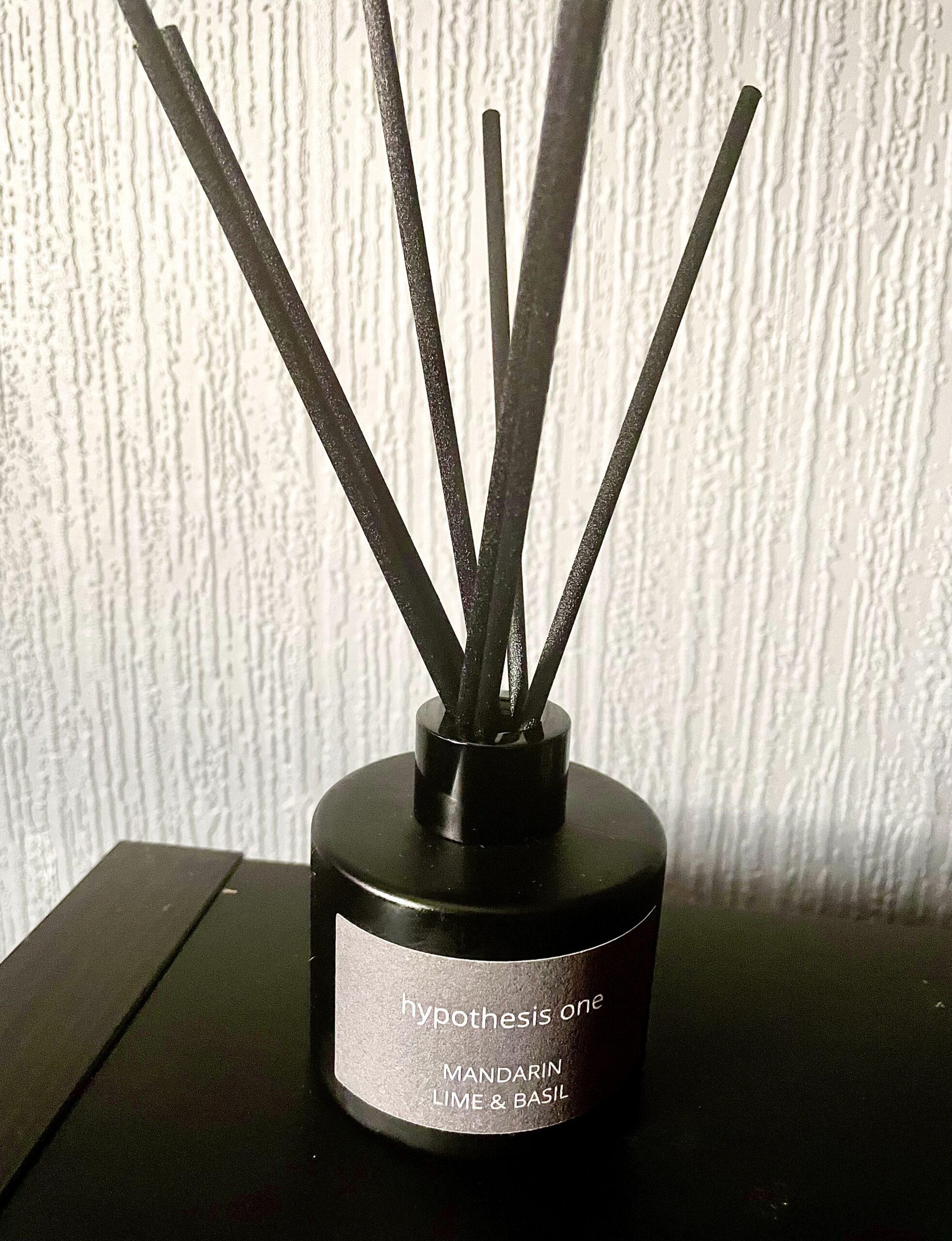 A short cylindrical black glass jar with a light grey label that has Hypothesis One Mandarin, Lime & Basil Intense Fragrance Diffuser written in bold white writing on it and some black wooden reeds coming out of the top of it on top of a black glossy wooden cabinet.