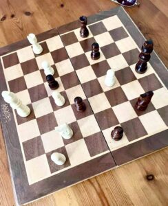 A large rectangular dark wooden and cream coloured checkered chessboard that has some dark wooden and white wooden chess pieces on top of it on a large light wooden table.