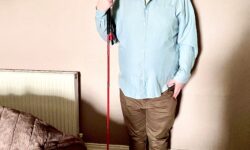 A tall bearded man wearing a pair of silver and black aviator Sunglasses with some light green lenses, a bright bluey green long sleeve oxford shirt, a bright green crew neck t-shirt, a pair of dark brown cord trousers and a pair of shiny red shoes holding a bright red cane stood against a light grey wall.