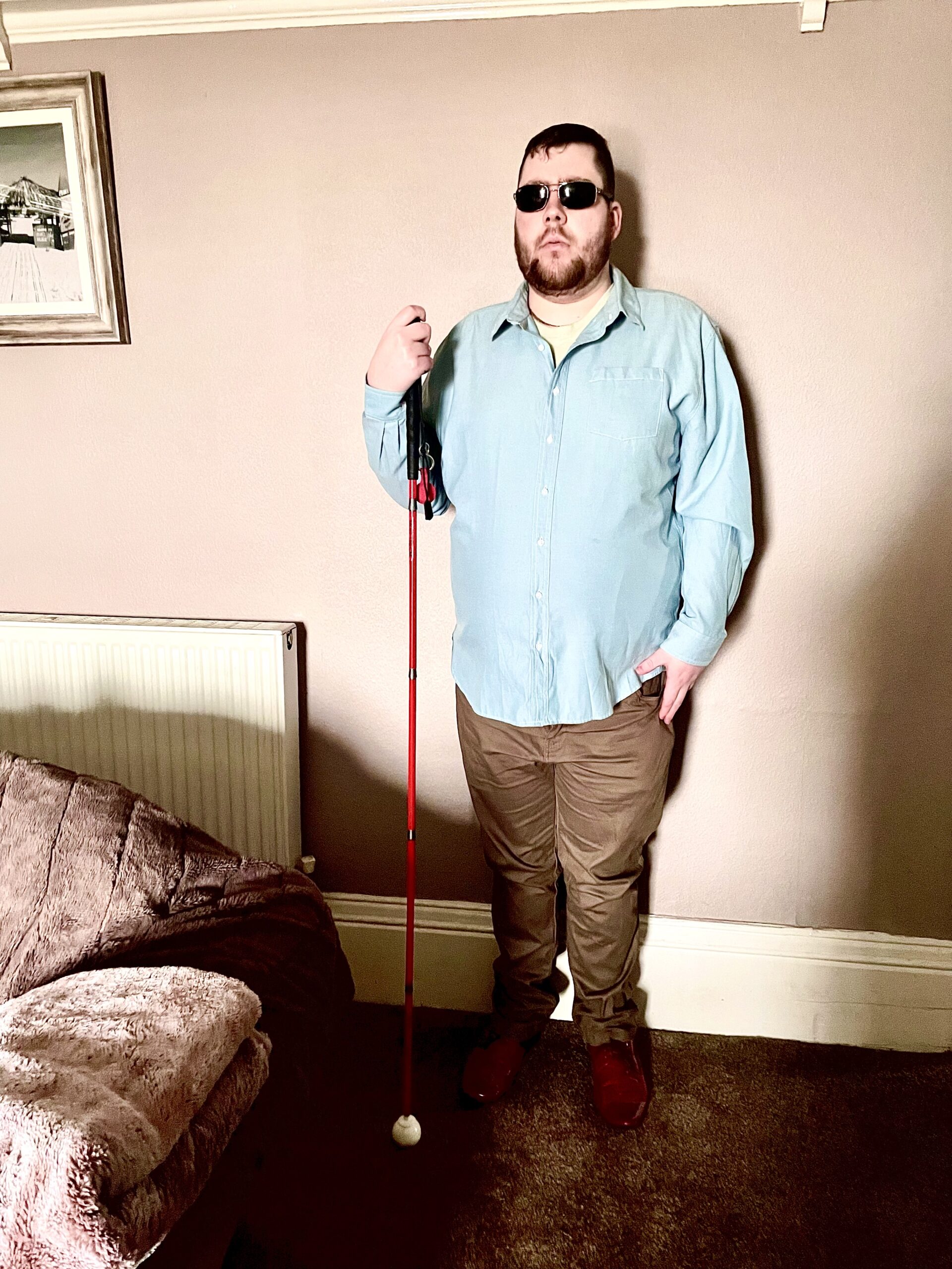 A tall bearded man wearing a pair of silver and black aviator Sunglasses with some light green lenses, a bright bluey green long sleeve oxford shirt, a bright green crew neck t-shirt, a pair of dark brown cord trousers and a pair of shiny red shoes holding a bright red cane stood against a light grey wall.
