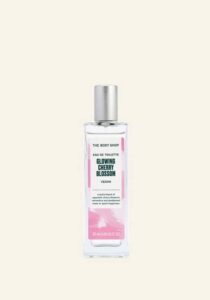 A tall rectangular clear glass bottle full of some clear liquid with a silver aluminium lid and a bright red label that has The Body Shop Glowing Cherry Blossom Eau De Toilette written in dark green writing and some pictures of some bright white cherry blossoms on it, on a white background.