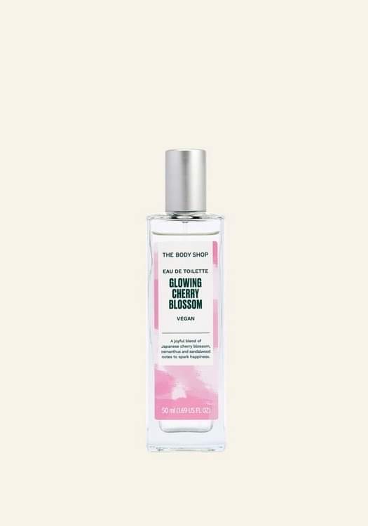 A tall rectangular clear glass bottle full of some clear liquid with a silver aluminium lid and a bright red label that has The Body Shop Glowing Cherry Blossom Eau De Toilette written in dark green writing and some pictures of some bright white cherry blossoms on it, on a white background.