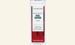 A tall rectangular clear glass bottle full of some clear liquid with a silver aluminium lid and a bright red label that has The Body Shop Rebel Rosebud Eau De Toilette written in dark green writing and some pictures of some dark red Roses on it, on a white background.
