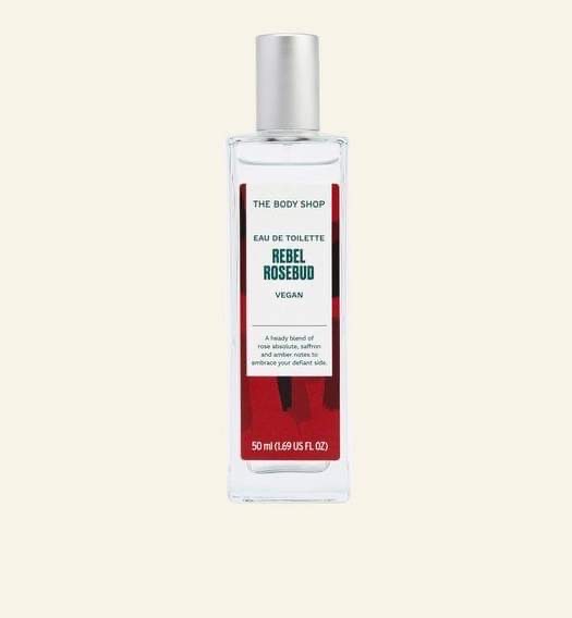 A tall rectangular clear glass bottle full of some clear liquid with a silver aluminium lid and a bright red label that has The Body Shop Rebel Rosebud Eau De Toilette written in dark green writing and some pictures of some dark red Roses on it, on a white background.