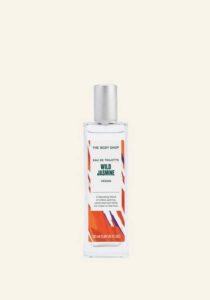 A tall rectangular clear glass bottle full of some clear liquid with a silver aluminium lid and a bright orange label that has The Body Shop Wild Jasmine Eau De Toilette written in dark green writing and some pictures of some bright green Jasmine Leaves on it, on a white background.
