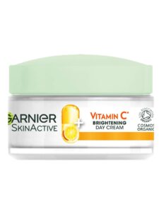 A short cylindrical white glass jar with a bright white plastic lid and a bright white label that has Garnier Vitamin C Brightening Day Cream written in bold black writing on it, on a white background.
