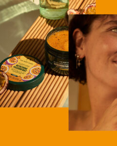A short cylindrical dark green plastic tub with a dark green plastic lid and a bright Yellow label that has The Body Shop Refreshing Passionfruit Body Scrub written in dark green writing and some pictures of some dark purple slices of Passionfruit on it, on a white background.
