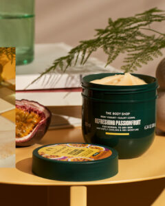 A short rounded rectangular dark green plastic tub with a dark green plastic lid and a bright Yellow label that has The Body Shop Refreshing Passionfruit Body Yoghurt written in dark green writing and some pictures of some dark purple slices of Passionfruit on it, on a white background.