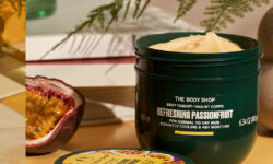 A short rounded rectangular dark green plastic tub with a dark green plastic lid and a bright Yellow label that has The Body Shop Refreshing Passionfruit Body Yoghurt written in dark green writing and some pictures of some dark purple slices of Passionfruit on it, on a white background.