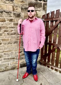 A tall bearded man wearing a pair of silver and black aviator sunglasses with some light green lenses, a bright pink long sleeve shirt, a dark red crew neck t-shirt, a pair of navy jeans and a pair of shiny bright red shoes holding a bright red cane stood against a dark wooden gate.