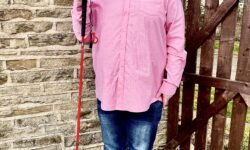 A tall bearded man wearing a pair of silver and black aviator sunglasses with some light green lenses, a bright pink long sleeve shirt, a dark red crew neck t-shirt, a pair of navy jeans and a pair of shiny bright red shoes holding a bright red cane stood against a dark wooden gate.