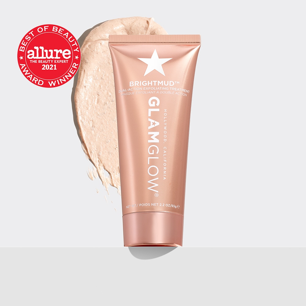 A short cylindrical rose gold Glass Jar with a rose gold lid that has GlamGlow BrightMud™ Dual-Exfoliating Treatment written in bold black writing and a picture of a silver star on it next to a rose gold diamond shaped cardboard box that has GlamGlow BrightMud™ Dual-Exfoliating Treatment written in white writing on it, on a white background.