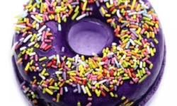 A large circular dark purple doughnut shaped bath bomb that has some various different coloured sprinkles embedded on top of it, , on a white background.