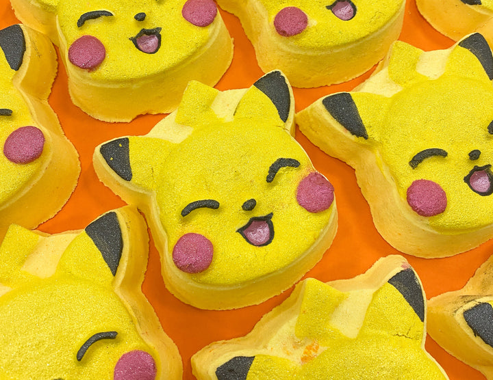 A large bright yellow squirrels head shaped bath bomb that has some black ears, some black eyes, a black mouth and some rosy red cheeks engraved on it, on a white background.