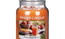 A tall cylindrical glass jar full of some bright peach wax with a label that has Yankee Candle written in white writing, Farm Fresh Peach written in black writing, and some pictures of some bright pink Peaches in a large circular bowl on it, on a white background.