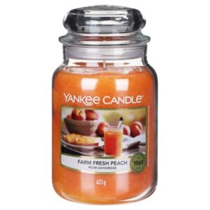 A tall cylindrical glass jar full of some bright peach wax with a label that has Yankee Candle written in white writing, Farm Fresh Peach written in black writing, and some pictures of some bright pink Peaches in a large circular bowl on it, on a white background.