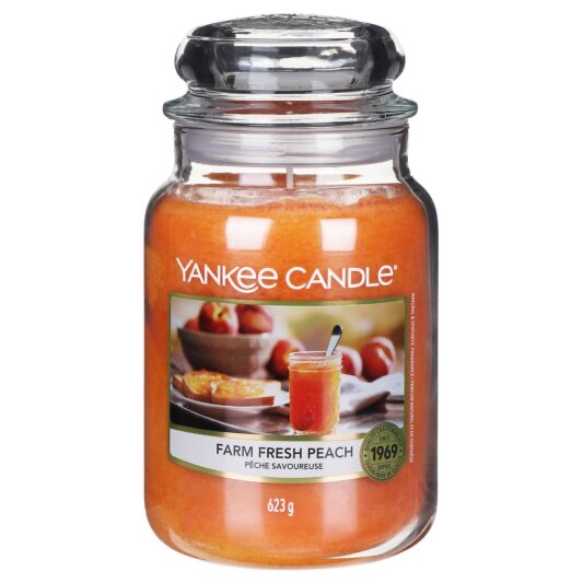 Picking Up Some Fresh Peaches From The Farm | Yankee Candle Farm Fresh Peach A tall cylindrical glass jar full of some bright peach wax with a label that has Yankee Candle written in white writing, Farm Fresh Peach written in black writing, and some pictures of some bright pink Peaches in a large circular bowl on it, on a white background.