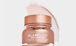 A short cylindrical clear plastic tub full of some bright gold liquid with a bright gold plastic lid that has GlamGlow BrightEyes Illuminating Anti-Fatigue Eye Cream written in bold white writing and a picture of a white star on it, on a white background.