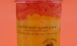 A medium cylindrical clear glass jar full of some bright red and yellow sugar scrub with a metallic silver lid that has H&L Bath Bombs Peach Sangria Fragranced Sugar Body Scrub written in bold black writing on it, on a white background.