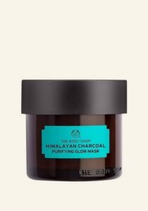 A short cylindrical black glass jar with a black plastic lid and a dark green label that has The Body Shop Himalayan Charcoal Purifying Glow Mask written in bold black writing on it, on a white background.