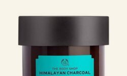 A short cylindrical black glass jar with a black plastic lid and a dark green label that has The Body Shop Himalayan Charcoal Purifying Glow Mask written in bold black writing on it, on a white background.