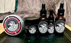 A short cylindrical brown glass jar with a black metallic lid and a gradient black and white label that has Arthur J Hawke Cherry Cola Beard Butter written in bold white writing and a picture of a black hawk on it, on a light wooden table..