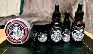 A short cylindrical brown glass jar with a black metallic lid and a gradient black and white label that has Arthur J Hawke Cherry Cola Beard Butter written in bold white writing and a picture of a black hawk on it, on a light wooden table..