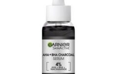 A short rounded cylindrical clear glass bottle full of some black liquid with a white rubber pipette lid and a silver label that has Garnier Skin Active 4% AHA + BHA & Niacinamide Charcoal Serum written in bold black writing on it, on a white background.