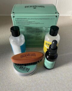 A short cylindrical brown glass bottle with a black rubber pipette lid and a bright green label that has Golden Beards Artic Beard Oil written in bold black writing and a picture of a beard on it next to a short cylindrical silver metallic tin with a silver metallic lid and a bright green label that has Golden Beards Artic Beard Balm written in bold black writing and a picture of a Beard on it, a short cylindrical black plastic bottle with a black plastic lid and a bright yellow label that has Golden Beards Beard Wash Shampoo written in bold black writing and a picture of a beard on it, a short cylindrical black plastic bottle with a black plastic lid and a bright blue label that has Golden Beards Beard Conditioner written in bold black writing and a picture of a Beard on it, a small oval light wooden beard brush with some black bristles that has Golden Beards engraved on it, and a large square bright red cardboard box that has Golden Beards Artic Beard Starter Set written in bold black writing on it, on a white background.