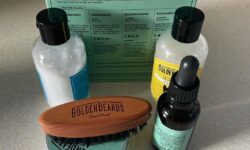 A short cylindrical brown glass bottle with a black rubber pipette lid and a bright green label that has Golden Beards Artic Beard Oil written in bold black writing and a picture of a beard on it next to a short cylindrical silver metallic tin with a silver metallic lid and a bright green label that has Golden Beards Artic Beard Balm written in bold black writing and a picture of a Beard on it, a short cylindrical black plastic bottle with a black plastic lid and a bright yellow label that has Golden Beards Beard Wash Shampoo written in bold black writing and a picture of a beard on it, a short cylindrical black plastic bottle with a black plastic lid and a bright blue label that has Golden Beards Beard Conditioner written in bold black writing and a picture of a Beard on it, a small oval light wooden beard brush with some black bristles that has Golden Beards engraved on it, and a large square bright red cardboard box that has Golden Beards Artic Beard Starter Set written in bold black writing on it, on a white background.