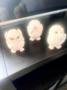 A small bright white owl figurine that has it’s wings over it’s ears next to a small bright white owl figurine that has it’s wings over it’s eyes, and a small bright white owl figurine that has it’s wings over it’s beak on a medium rectangular black wooden cabinet.