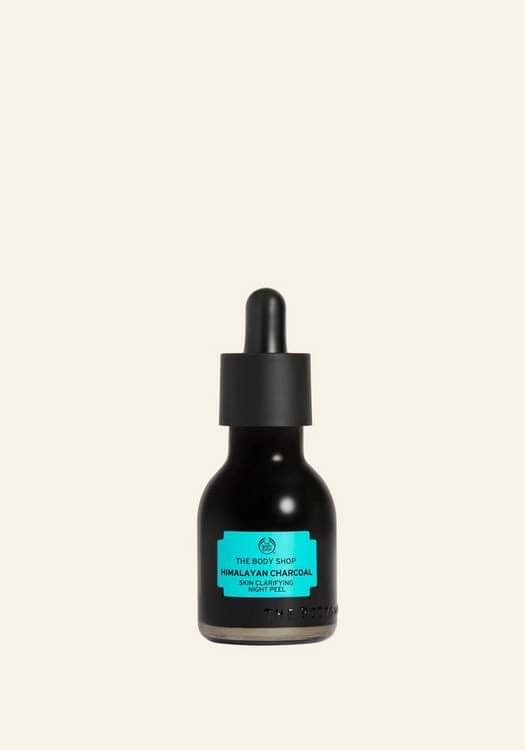 A short cylindrical clear glass bottle full of some black liquid with a black rubber pipette lid and a light blue label that has The Body Shop Himalayan Charcoal Skin Clarifying Night Peel written in bold black writing on it, on a white background.