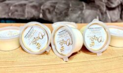 A collection of six bright green cylindrical blocks of wax inside some small cylindrical Plastic Tubs that all have white labels that has Dayas Essence written in bright gold writing on them, on a white background.