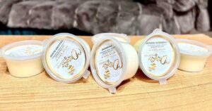 A collection of six bright green cylindrical blocks of wax inside some small cylindrical Plastic Tubs that all have white labels that has Dayas Essence written in bright gold writing on them, on a white background.