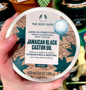 A short cylindrical silver metallic tin with a silver metallic lid and a white label that has The Body Shop Jamaican Black Castor Oil Leave-In Conditioner written in dark green writing on it on a light wooden shelf.