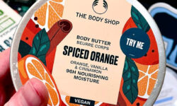 A short cylindrical silver metallic tin full of some creamy white body butter with a silver metallic lid and a white label that has The Body Shop Spiced Orange Body Butter written in dark green writing and some pictures of some large spherical Oranges on it, on a white background.